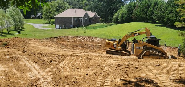 Excavator and skid steer cutting a pond basin out of a lawn