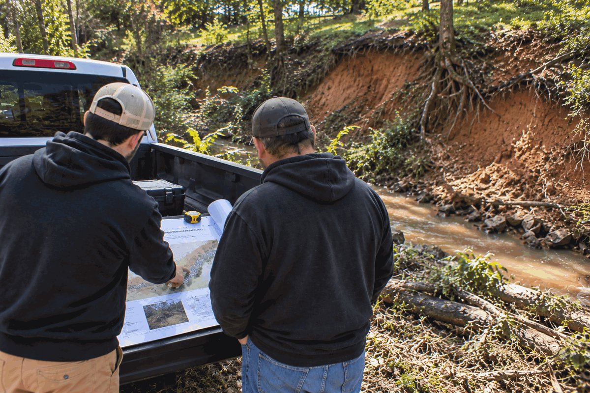 Two crew members going over a bank repair plan on a pickup tailgate beside an eroded creek