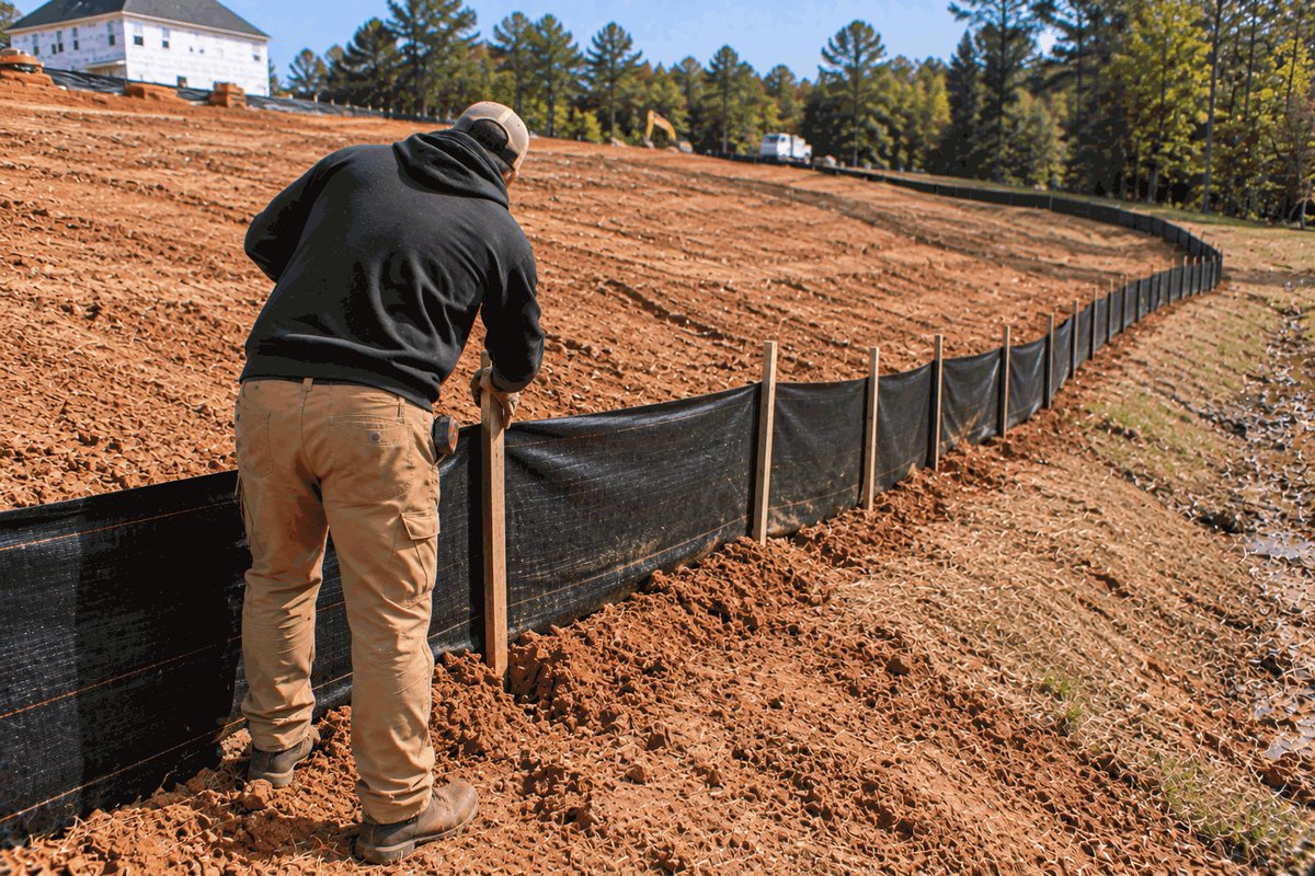 Worker setting a wooden stake in a line of black silt fence trenched along the bottom of a graded slope