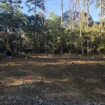 Construction site in a forest clearing with a yellow excavator on the left and cleared dirt ground extending to a tree line in the background